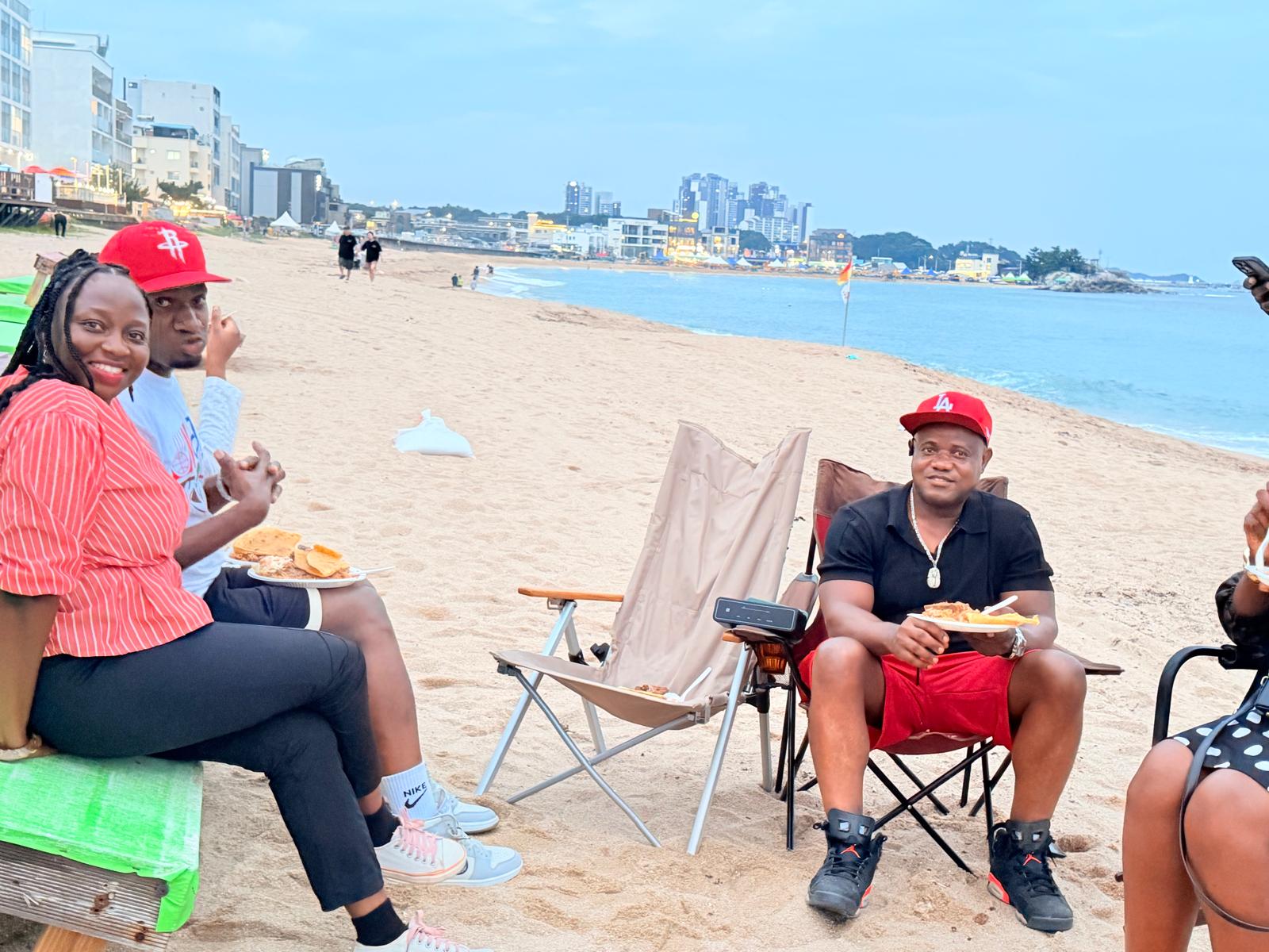 USKA members eating on folding chairs at Bongpo Beach, with the Gangwon coastline and town behind them